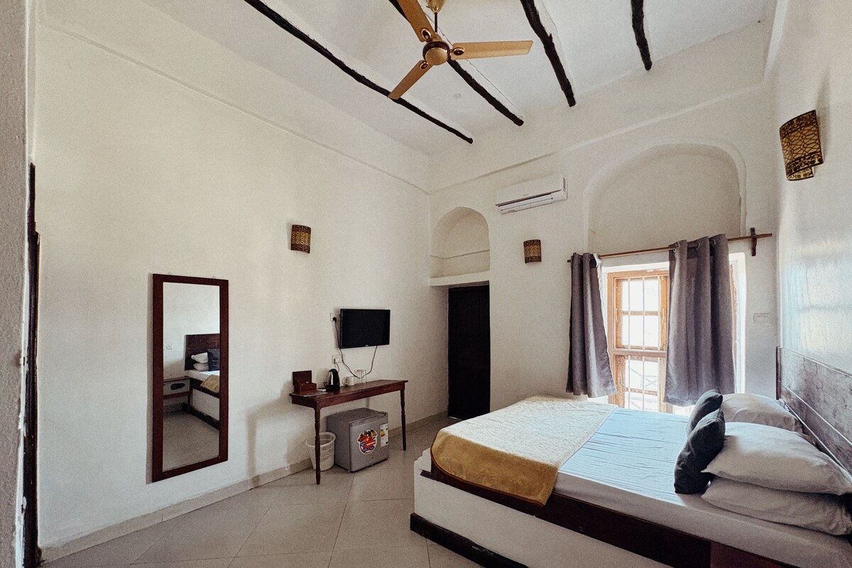 A bedroom with a bed, a desk, a television, and a ceiling fan.