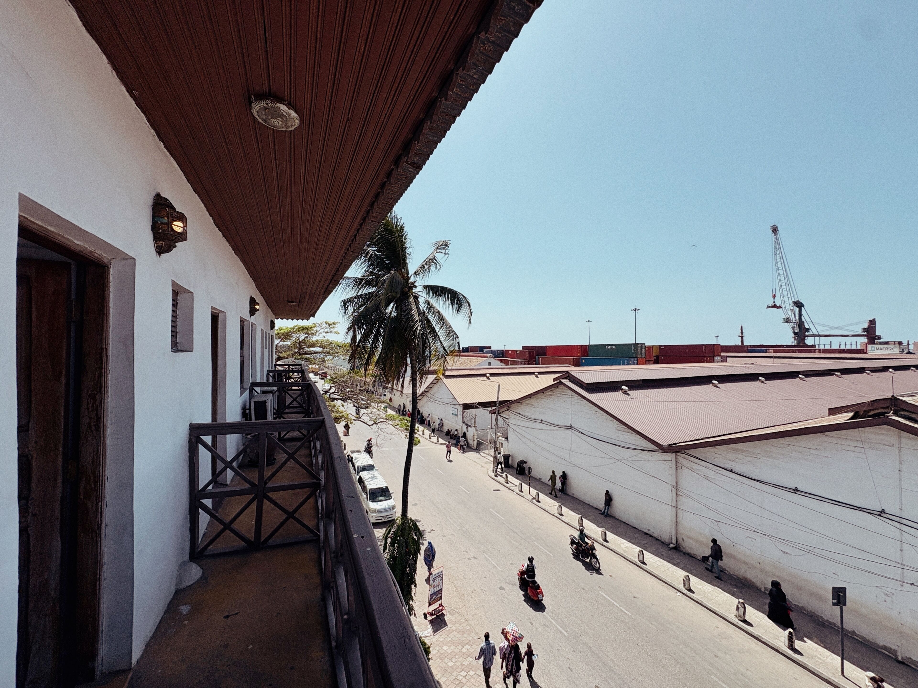 Un balcone con vista su una strada animata, palme e container in lontananza.