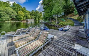Una terraza de madera con sillones de descanso y un bote amarrado a un lago, rodeado de árboles y una casa.