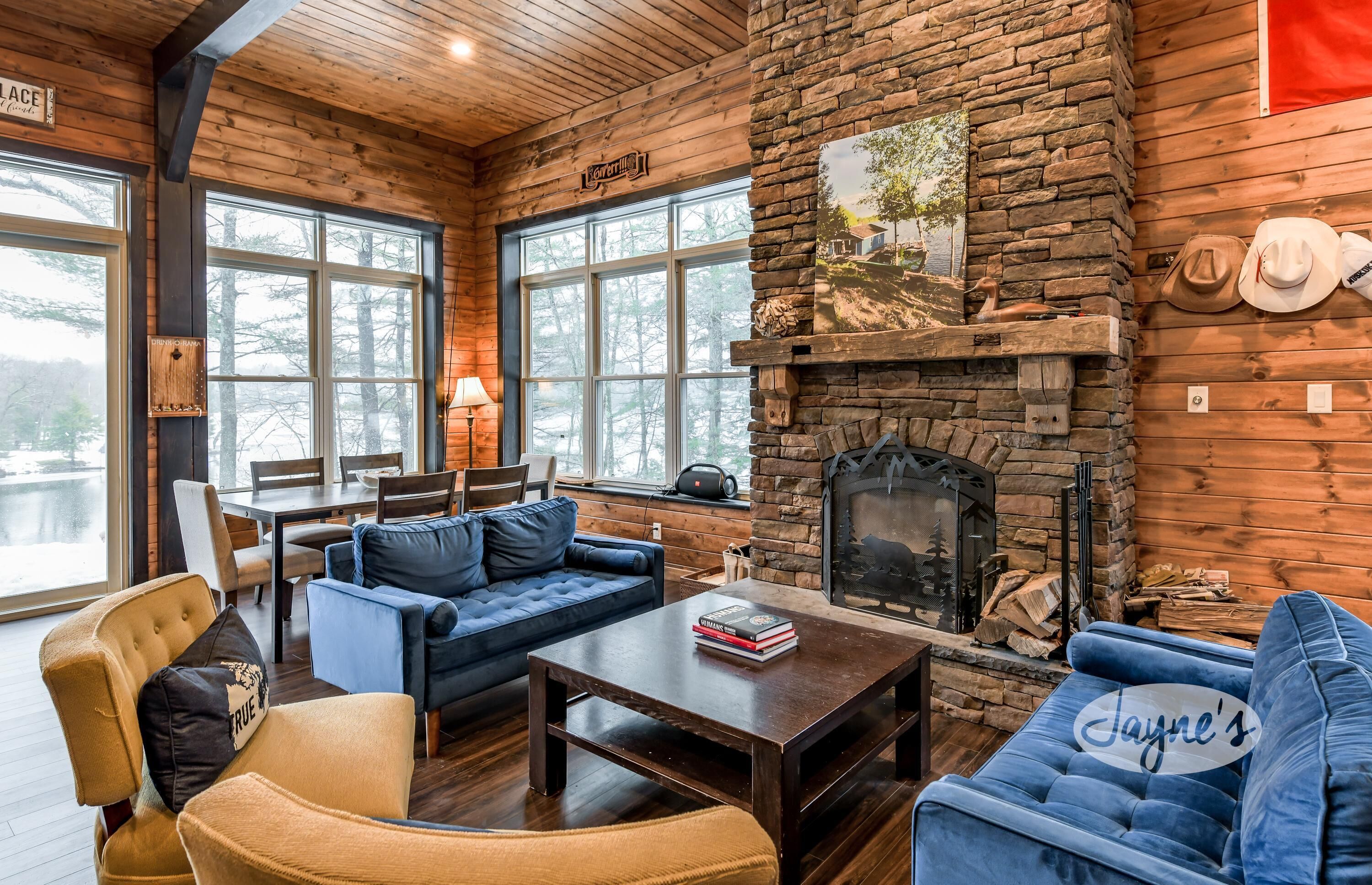 A cozy cabin interior with a stone fireplace, wooden walls, and large windows offering a view of the outdoors.