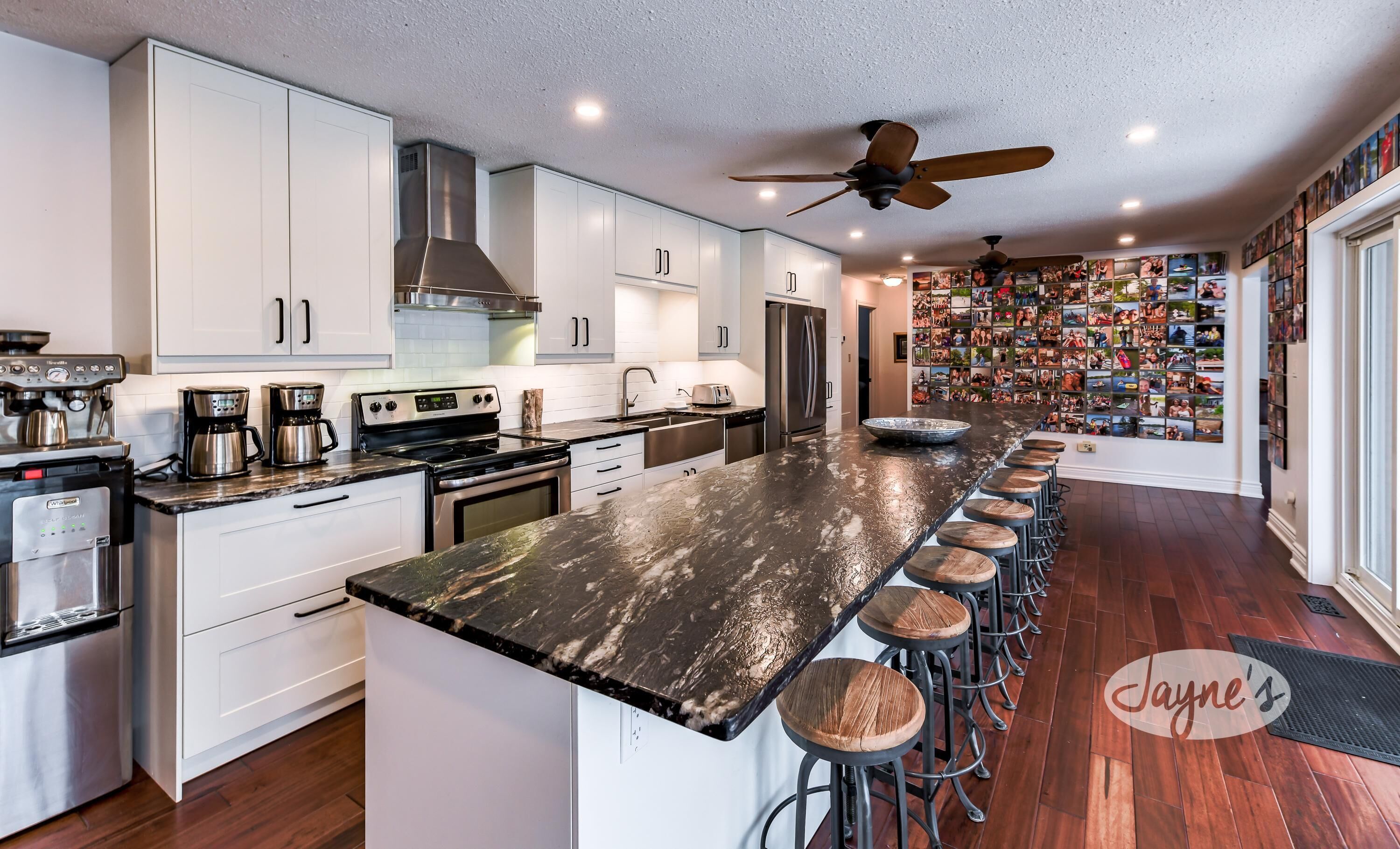 A modern kitchen with a central island, bar stools, stainless steel appliances, and a wall of photos.