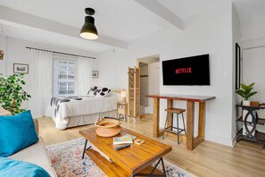 A modern living room with a bed, a coffee table, a TV, and a bookshelf. - Sunny Art Deco Studio – Walk to Conv Ctr & DTLB (Long Beach)