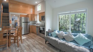 Una cocina moderna con armarios de madera, una zona de comedor con mesa y sillas, un sofá con cojines, y un ventanal con vistas a un bosque.