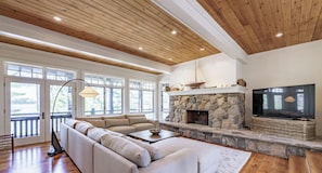 A living room with a stone fireplace, a large sofa, a television, and a wooden ceiling. - Traditional 6 bed 5 bath cottage on Lake Joseph with boathouse & pickleball! (MacTier)