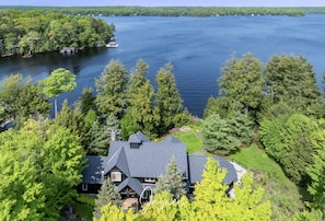 Une grande maison avec un toit en pente, entourée d’une forêt dense et bordée d’un lac.