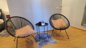 Ein Zimmer mit zwei schwarzen Stuhlen mit Drahtgestell und flauschigen Kissen, einem kleinen runden Tisch mit einer Pflanze und einem grauen, flauschigen Teppich auf Holzdielen.