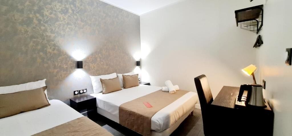 A hotel room with two beds, a desk, and a chair. There are wall-mounted lamps and a small shelf.