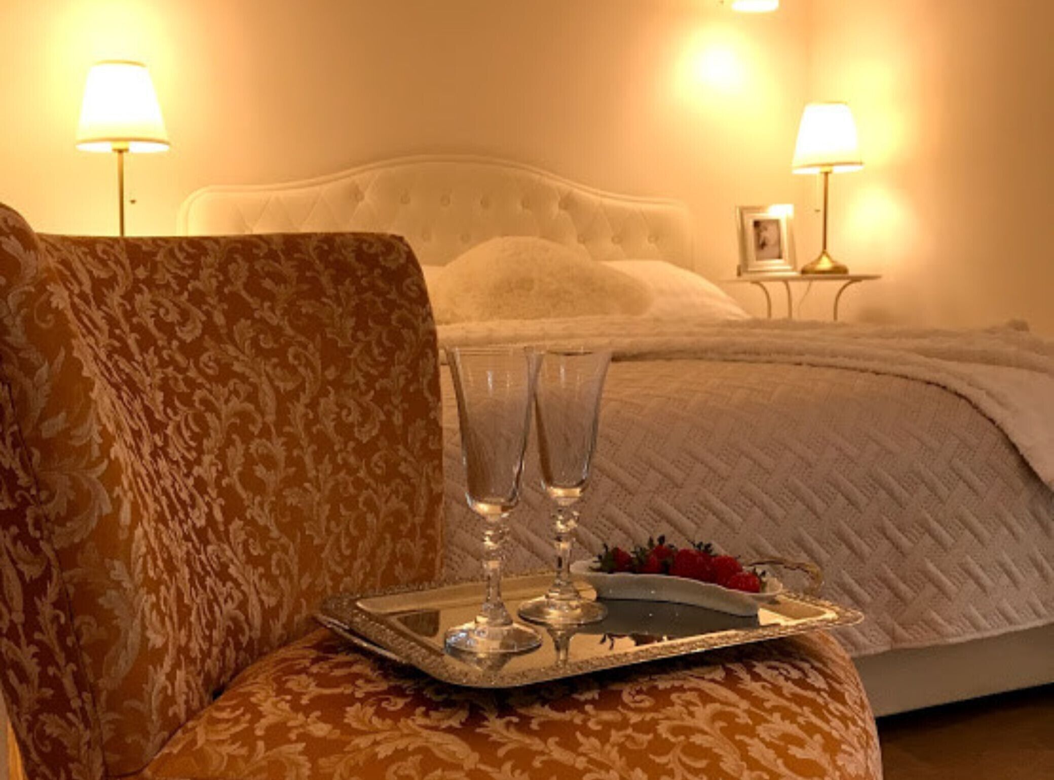 A bed with a white headboard, two bedside lamps, and a tray with two glasses and strawberries.