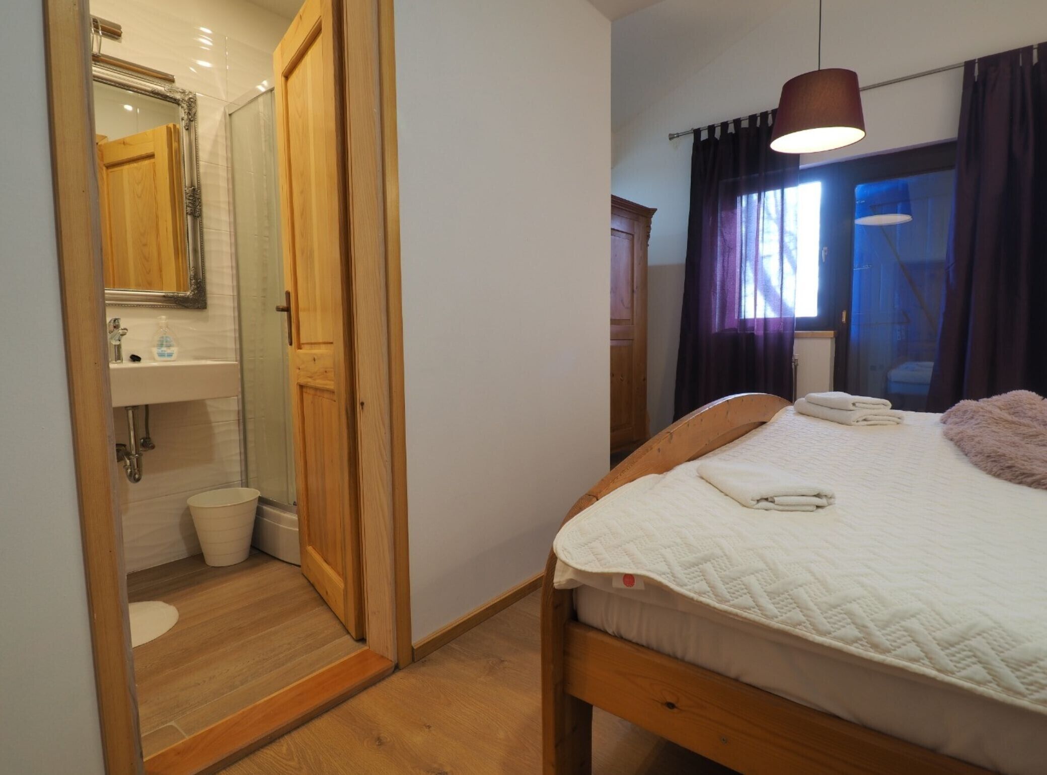 A bedroom with a wooden bed, a bedside table, a mirror, a window with curtains, and a bathroom with a sink and toilet.