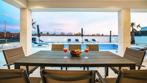 Ein Essbereich im Freien mit Blick auf einen Pool, ein Tisch mit Stühlen sowie eine Schüssel mit Obst und Getränken.