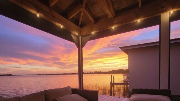 A covered outdoor seating area with a view of the sunset over water, wooden beams, and string lights. - Luxury Home on Dora Lake – 2 Min to Downtown Mount Dora (Mount Dora)