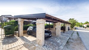 Ein überdachter Carport mit Steinpfeilern und einem Ziegeldach, neben einer Steinmauer und einem Metallgitter.