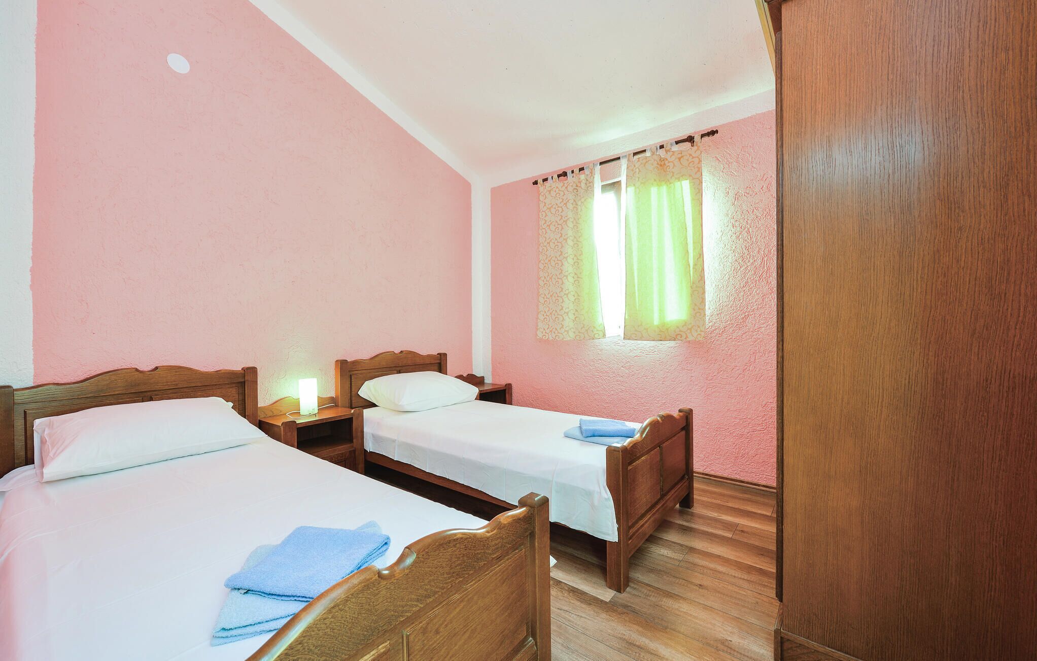 A room with two wooden beds, a nightstand with a lamp, a window with curtains, and a wooden wardrobe.