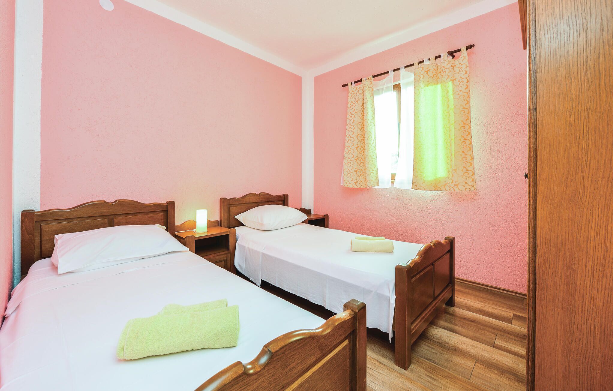 A room with two single beds, wooden headboards, a nightstand with a lamp, and a window with patterned curtains.