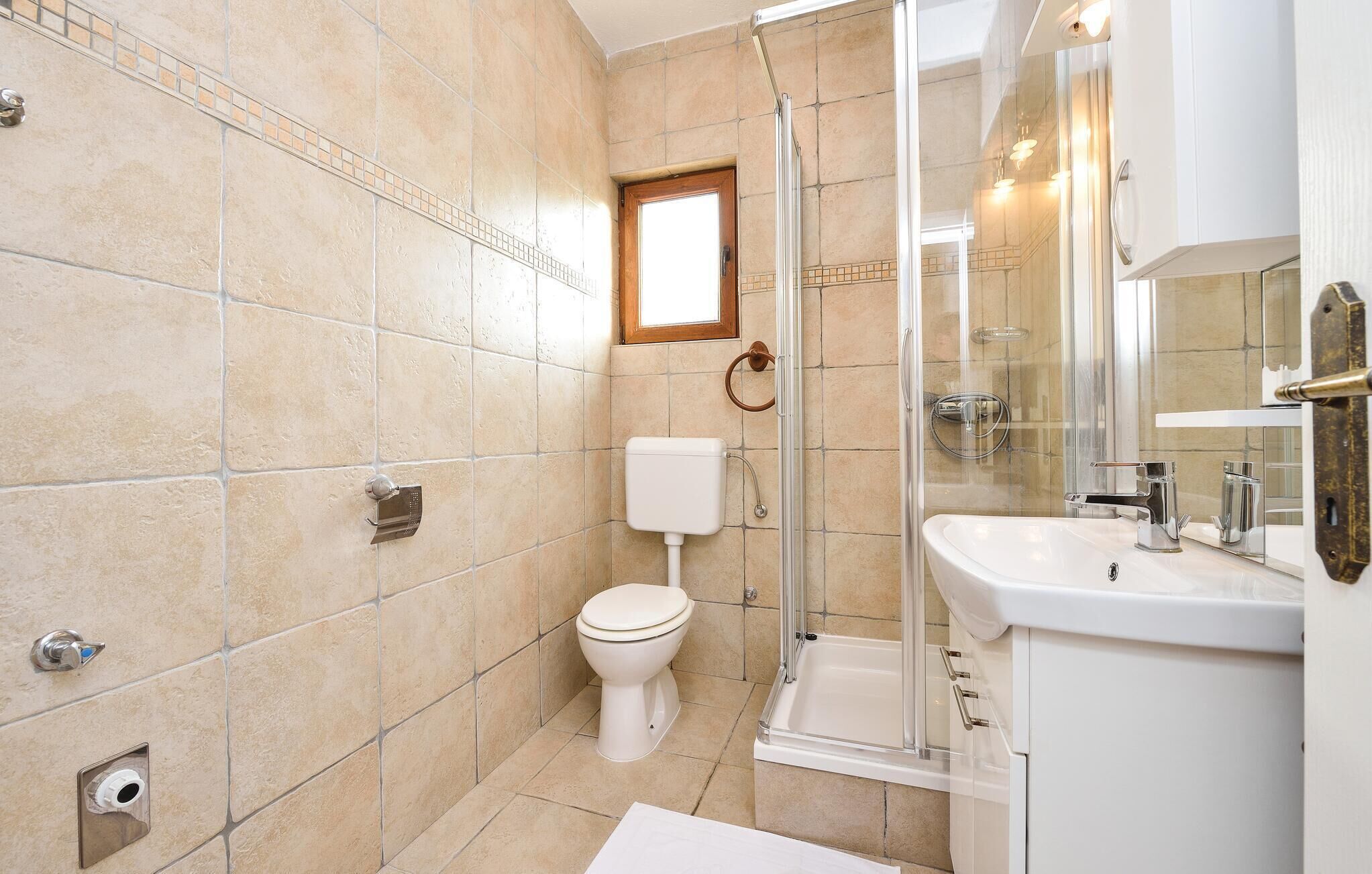 A modern bathroom with beige tiles, a glass-enclosed shower, a white toilet, and a white sink with a mirror above it.
