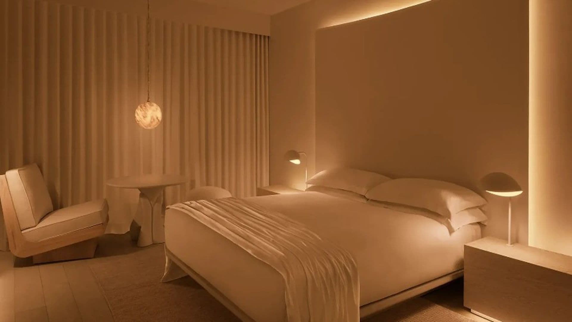 Een moderne hotelkamer met een groot bed, een klein rond tafeltje, een stoel en een nachtkastje met een lamp.