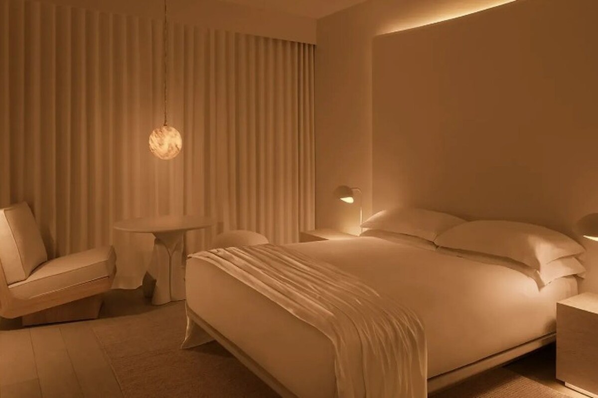 A modern hotel room with a large bed, a small round table, a chair, and a nightstand with a lamp.