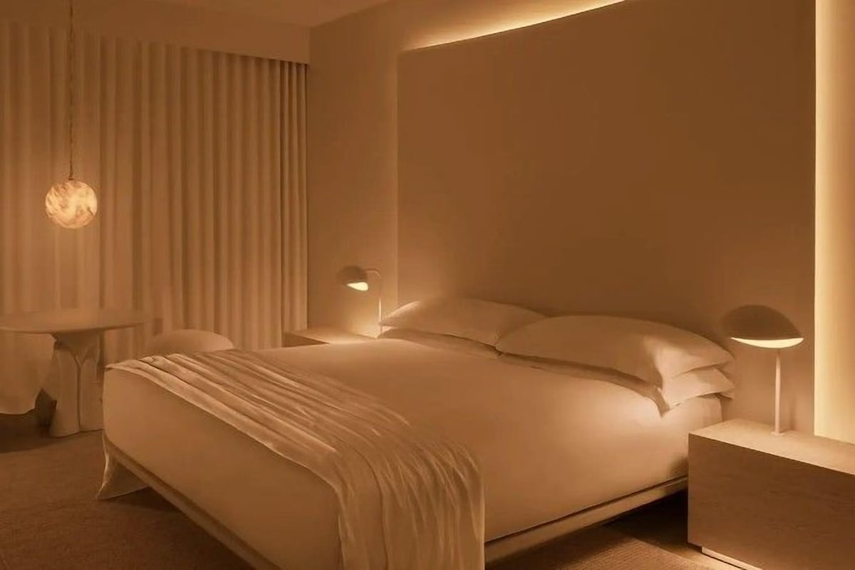 A modern hotel room with a large bed, a bedside table, a chair, and a wall-mounted lamp.