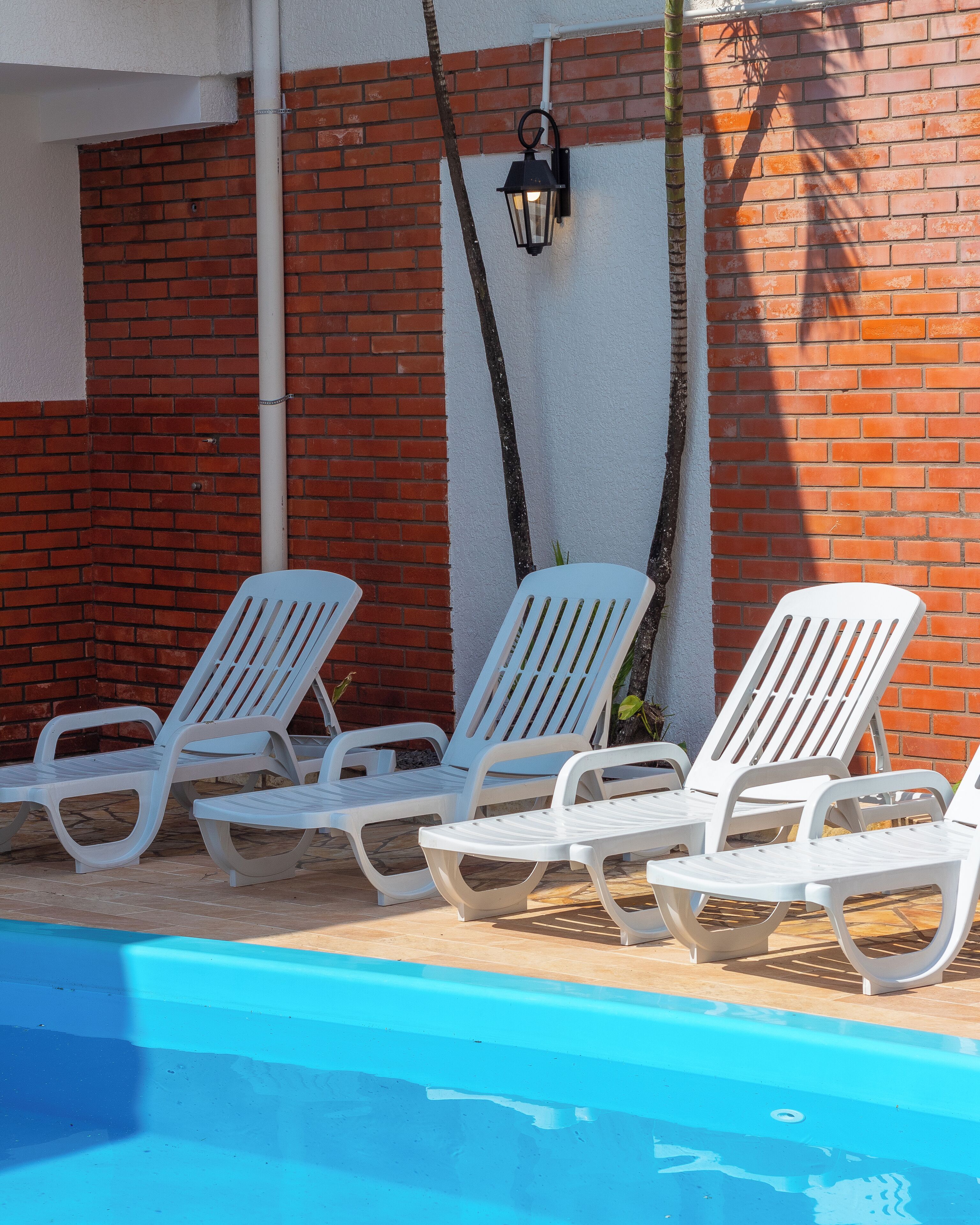 Zona junto a la piscina con sillas reclinables blancas y un aplique negro fijado a la pared.