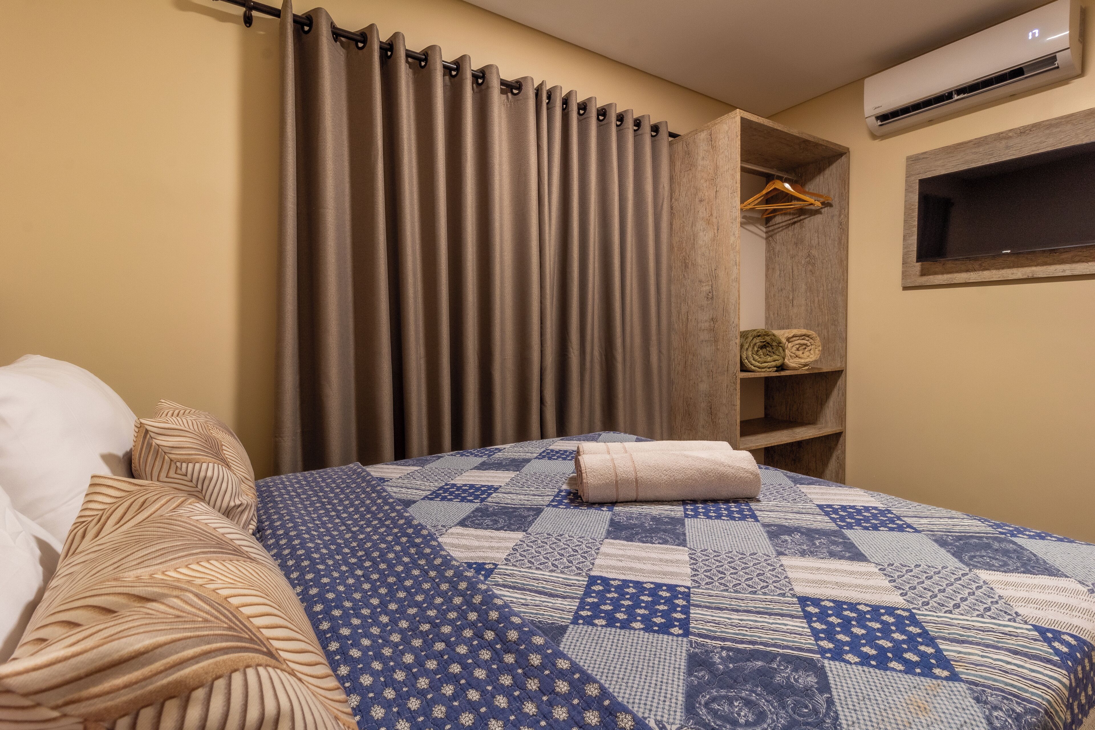 A neatly made bed with a blue and white quilt, a wooden shelf with folded towels, and a window with a wooden frame.