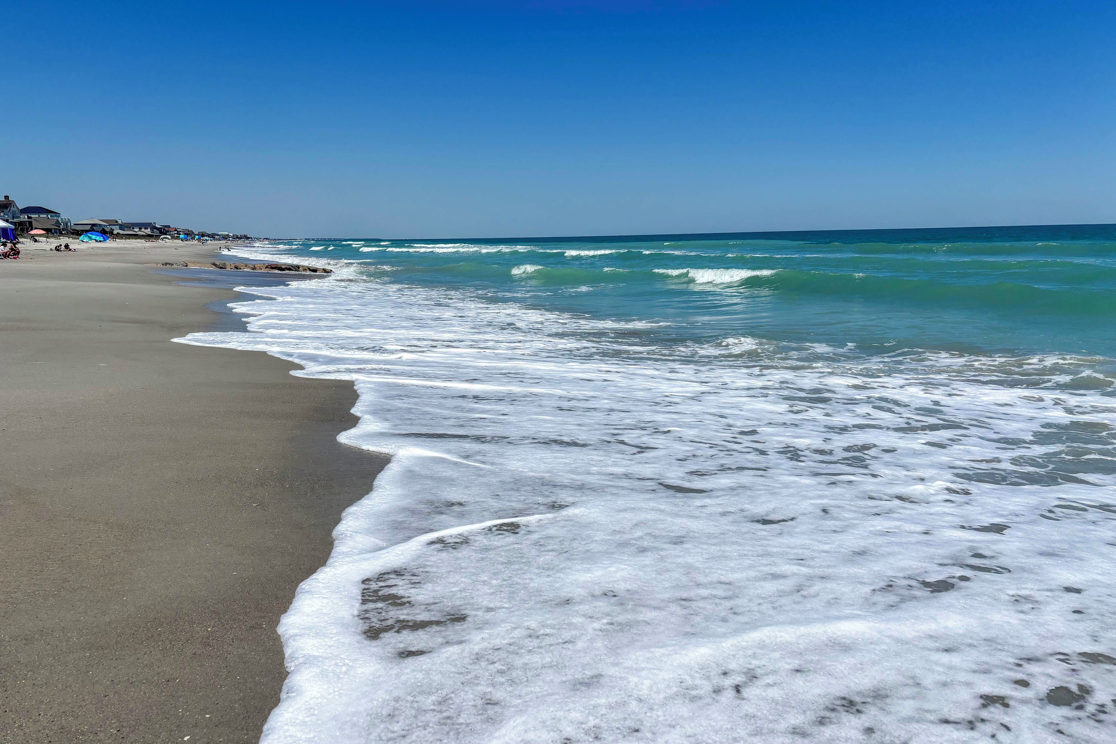 A beach with waves lapping onto the shore, a clear sky, and buildings in the background.