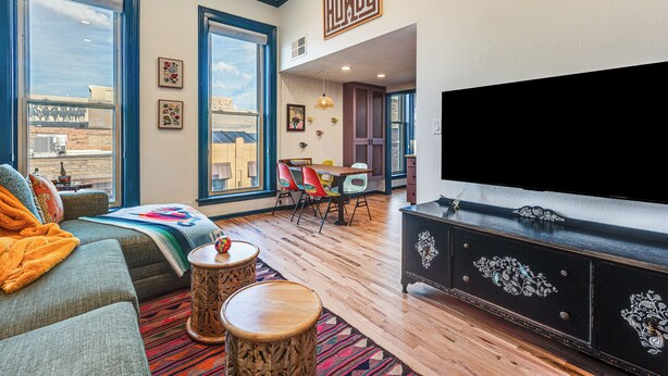 Een ruime woonkamer met een grote televisie, een bank, een eethoek met kleurrijke stoelen en grote ramen met uitzicht op de stad.
