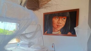 Ett sovrum med en säng täckt av ett vitt sänghimmel, ett litet träbord vid sängen med en lampa, en grön hopfällbar stol och en tavla på väggen.