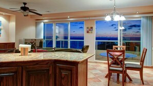 Eine moderne Küche mit einer großen Kochinsel, einem Essbereich mit Glastisch und Blick auf den Sonnenuntergang über dem Meer.