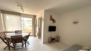 A room with a dining table, chairs, a bed with pillows, a TV, and a balcony with curtains. - Apartment for 4 with parking near beach (Argelès-sur-Mer)