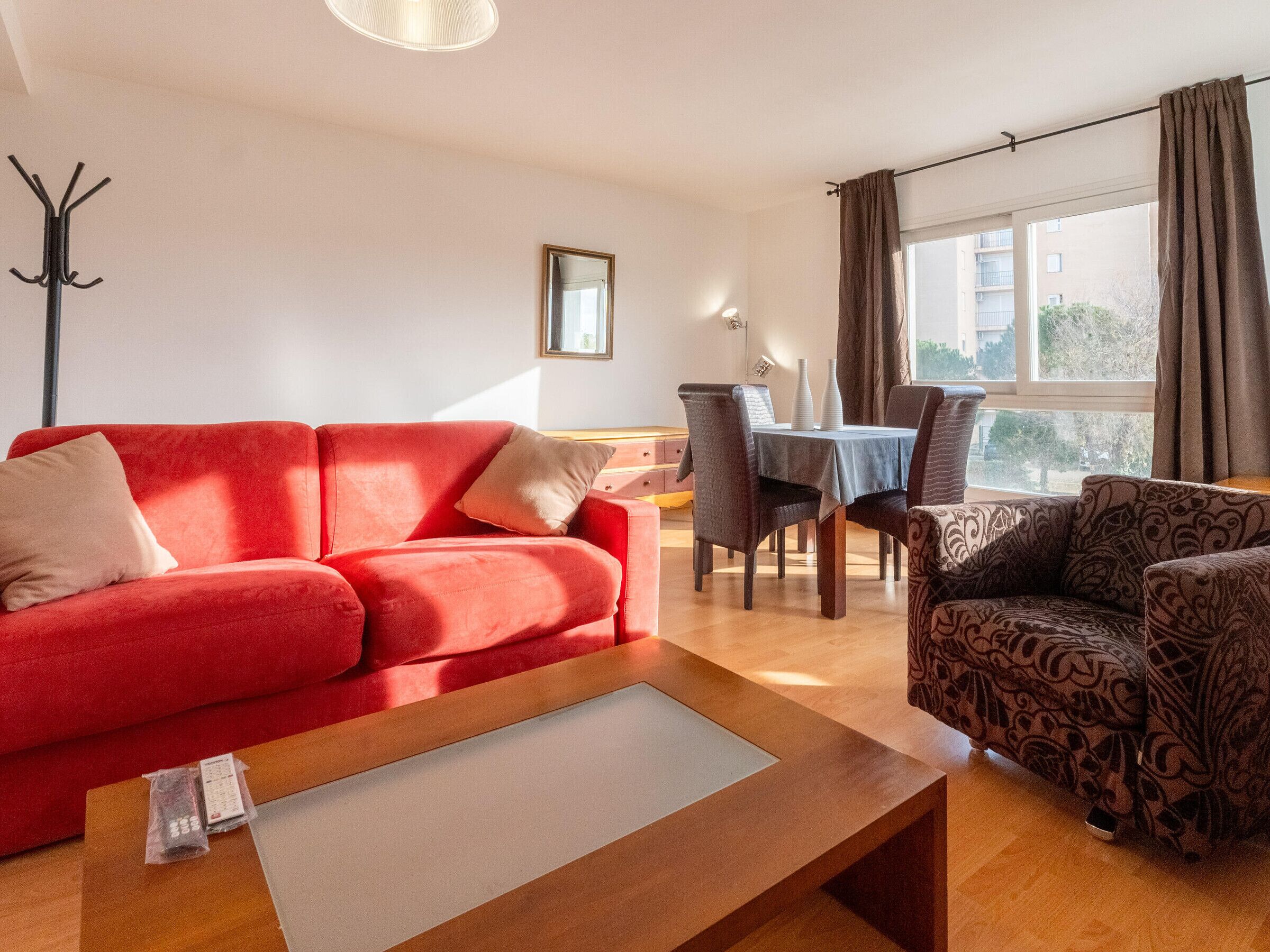 A living room with a red sofa, patterned armchair, and a wooden coffee table. There is a dining table with chairs and a window with curtains.