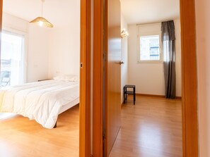 Ein Schlafzimmer mit Bett, einem kleinen Hocker, einem Fenster mit Vorhängen und einer Tür.