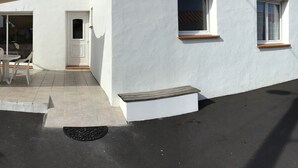 Ein weißes Gebäude mit einer gefliesten Terrasse, einer Tür und zwei Fenstern mit Holzbrettern.