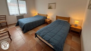 Ein Zimmer mit zwei Betten, jeweils mit einem hölzernen Kopfteil und einem Nachttisch mit einer Lampe. Ein Sessel und ein Fenster mit Jalousien sind ebenfalls vorhanden.