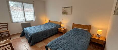 A room with two beds, each with a wooden headboard and a bedside table with a lamp. There is a chair and a window with blinds.