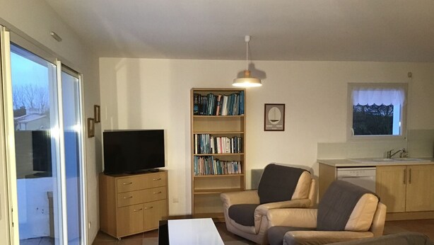 Ein Wohnzimmer mit Sofa, Sessel, Fernseher, Bücherregal und einer Küchenzeile mit Fenster.