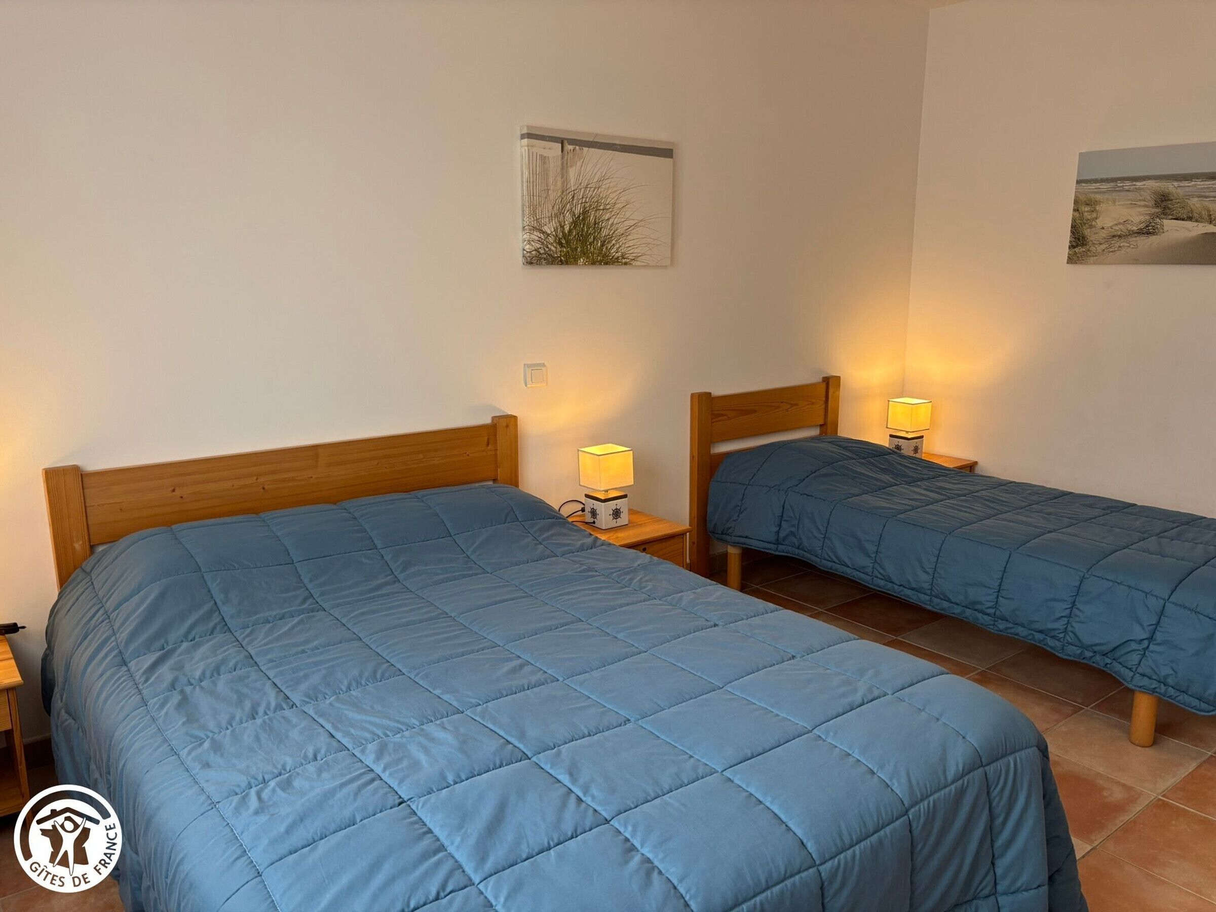 Une chambre avec deux lits, chacun doté d’une tête de lit en bois et d’une couvre-lit matelassée bleue. On y trouve des tables de chevet avec des lampes, ainsi qu’un tableau encadré au mur.