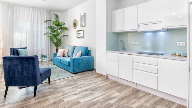 Eine moderne Wohnung mit Wohnzimmer, einem blauen Sofa, Essbereich und einer Küche mit weißen Schränken.