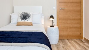 A neatly made bed with a dark blue blanket, white pillows, and a decorative pillow with a palm leaf design. There is a small bedside table with a lamp and a white vase. A wooden door is visible in the background. - Spacious 5-Bedroom Apartment near Guadalmedina River, Málaga (Málaga)