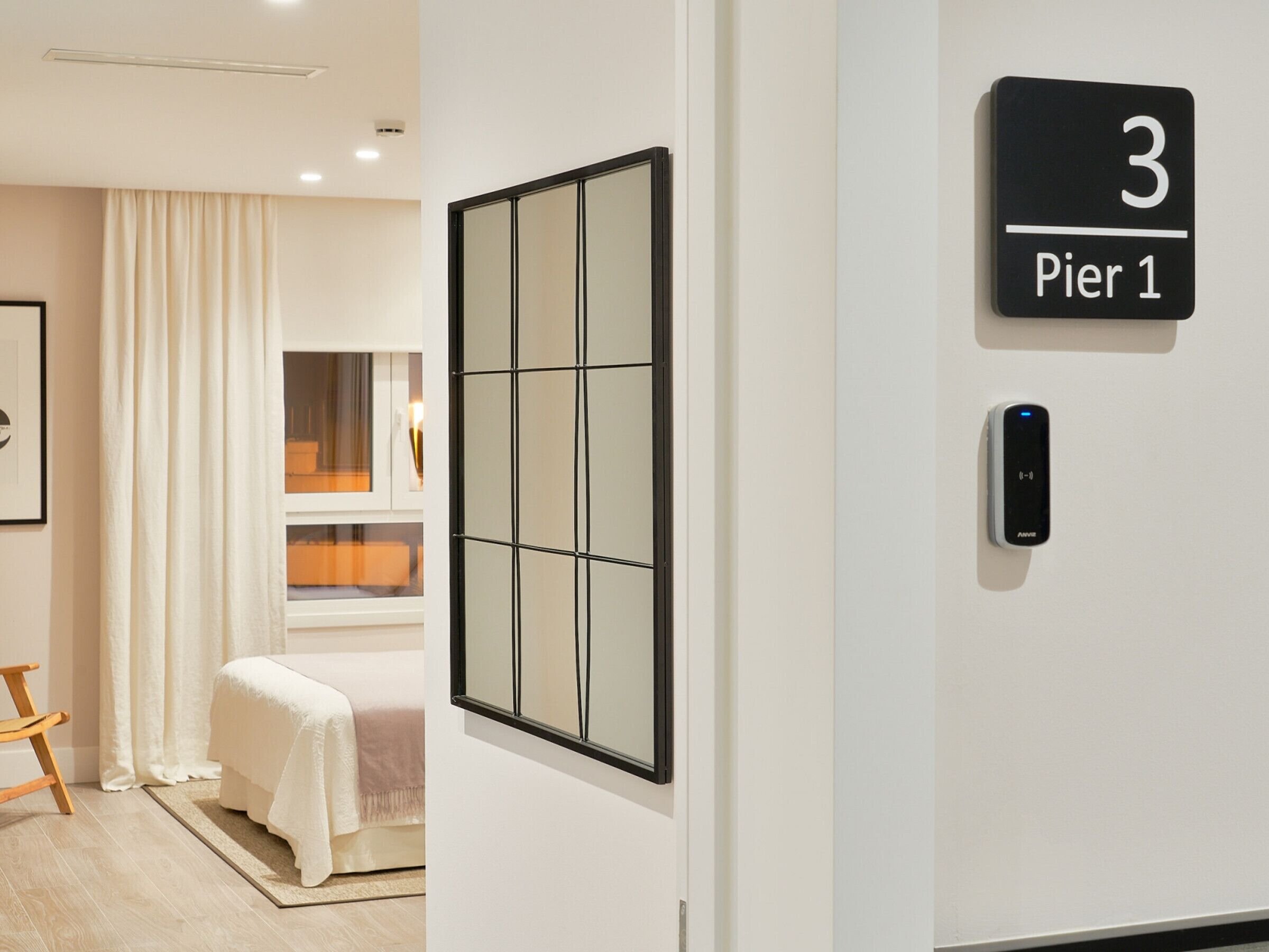 Ein Hotelzimmer mit Bett, Fenster mit Vorhängen, Nachttisch, fest montiertem Bedienfeld und einem gerahmten Kunstwerk.