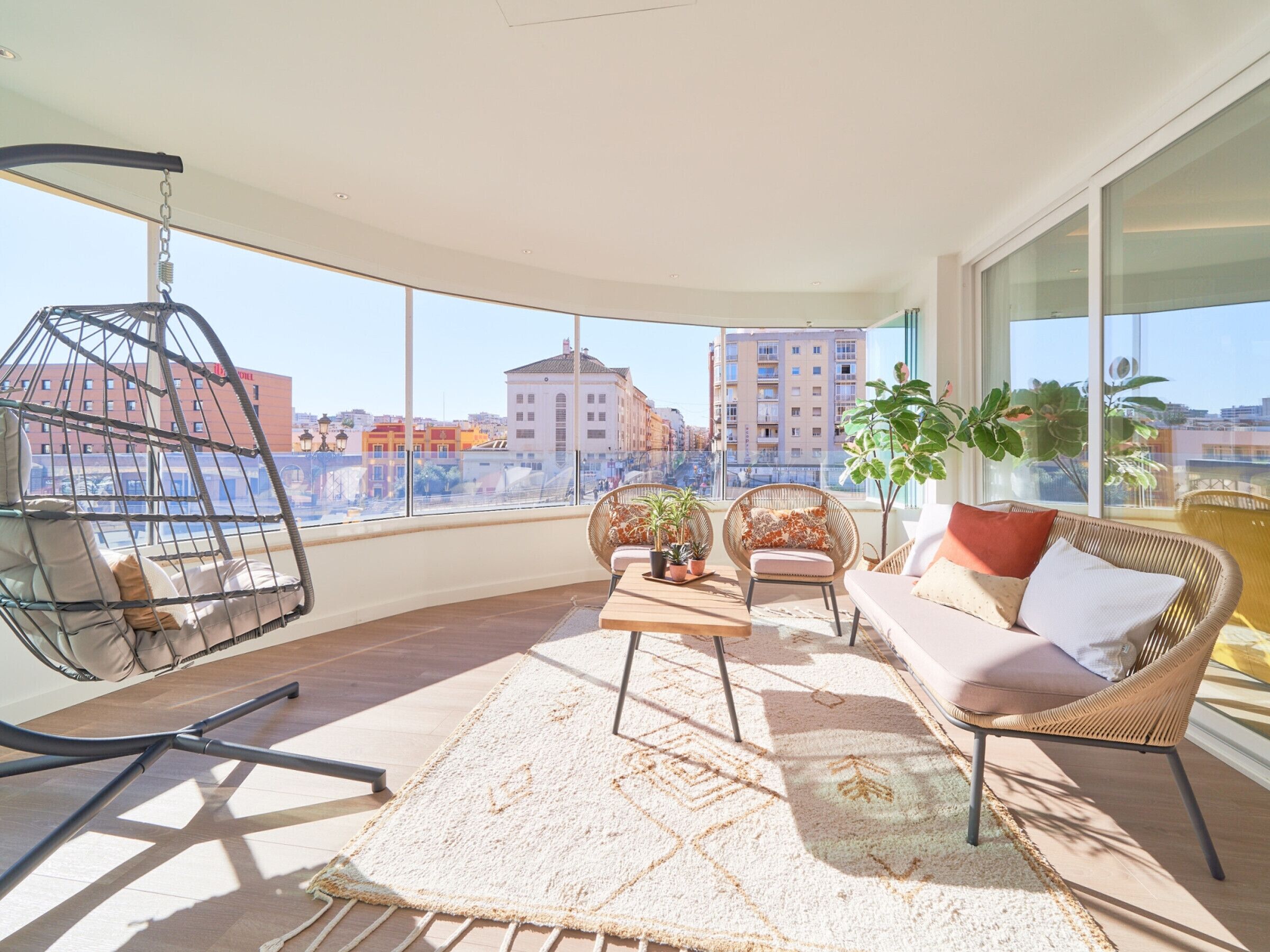 A spacious balcony with a hanging chair, a sofa, and a coffee table, featuring a city view.