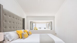 Ein Schlafzimmer mit einem großen Bett, einem grauen, gepolsterten Kopfteil und einem Fenster mit Vorhängen.