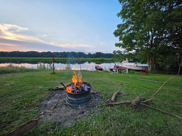 A campfire in a grassy area with a lake and boats in the background.