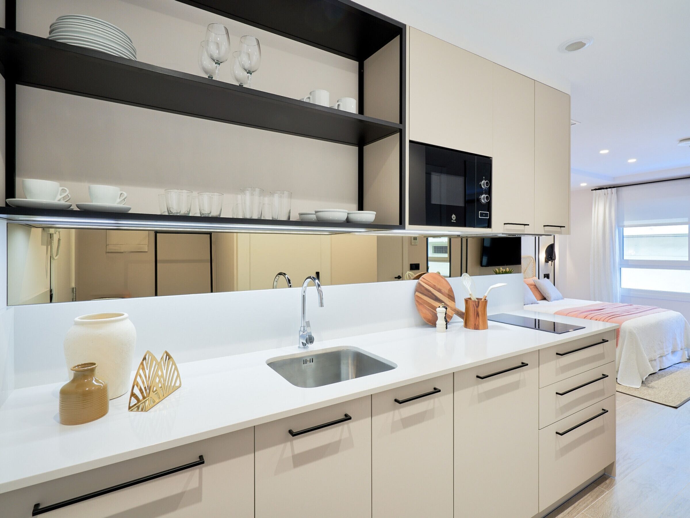 A modern kitchen with a white countertop, stainless steel sink, and built-in appliances. There are cabinets with black handles and a large mirror above the countertop.