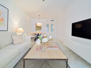 A modern living room with a sofa, coffee table, TV, and a dining area with chairs and a table. - Charming 3-Bed Apartment with Pool & Parking in Exclusive Miraflores del Palo, Málaga (Málaga)
