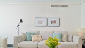 Een moderne woonkamer met een beige bank, een ronde salontafel en een vaas met gele bloemen. Boven de bank hangen twee ingelijste foto’s.