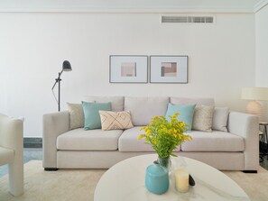 Ein modernes Wohnzimmer mit einem beigefarbenen Sofa, einem runden Couchtisch und einer Vase mit gelben Blumen. Darüber hängen zwei gerahmte Bilder an der Wand.