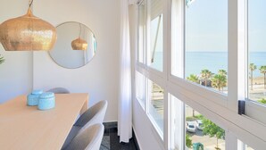 Een kamer met een houten tafel, twee stoelen, een hanglamp, een ronde spiegel en uitzicht op het strand door grote ramen.