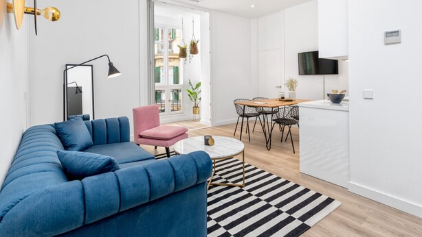 Ein modernes Wohnzimmer mit einem blauen Sofa, einem pinken Sessel, einem Essbereich mit Tisch und Stühlen sowie einer Küche mit Fernseher.