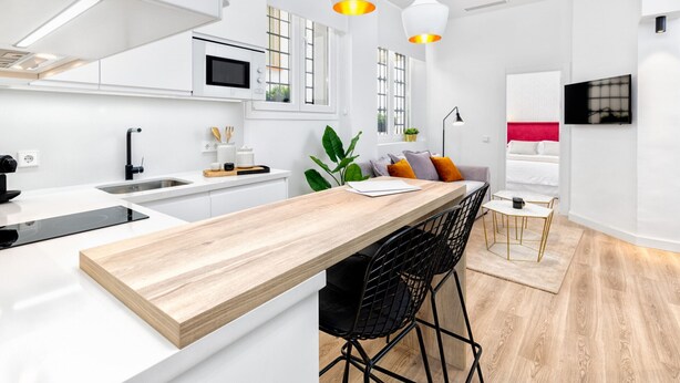 Una cocina moderna con armarios blancos, una isla de madera, taburetes negros y luces colgantes.