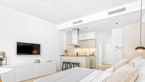 Un dormitorio moderno con decoración en blanco, una cama grande, un televisor y una kitchenette al fondo.